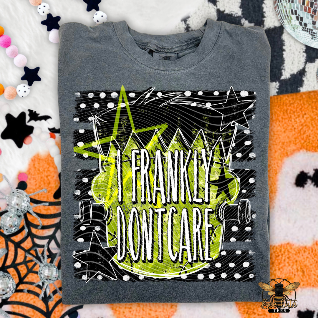A folded gray crewneck t-shirt with a graphic print reading 'I FRANKLY DONT CARE' in white over a lime green splatter shape, surrounded by black spiderwebs and white dots. The shirt is laid flat on a patterned orange, white, and black blanket with Halloween-themed props like spiderwebs, stars, and a disco ball.