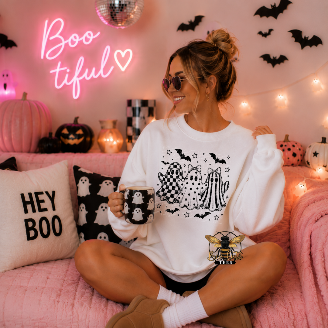 A woman sits cross-legged on a pink couch wearing a white crewneck sweatshirt with a black and white Halloween ghost graphic. The room is decorated for Halloween with a pink neon 'Boo tiful' sign, string lights, pumpkins, and black bat cutouts on the wall.