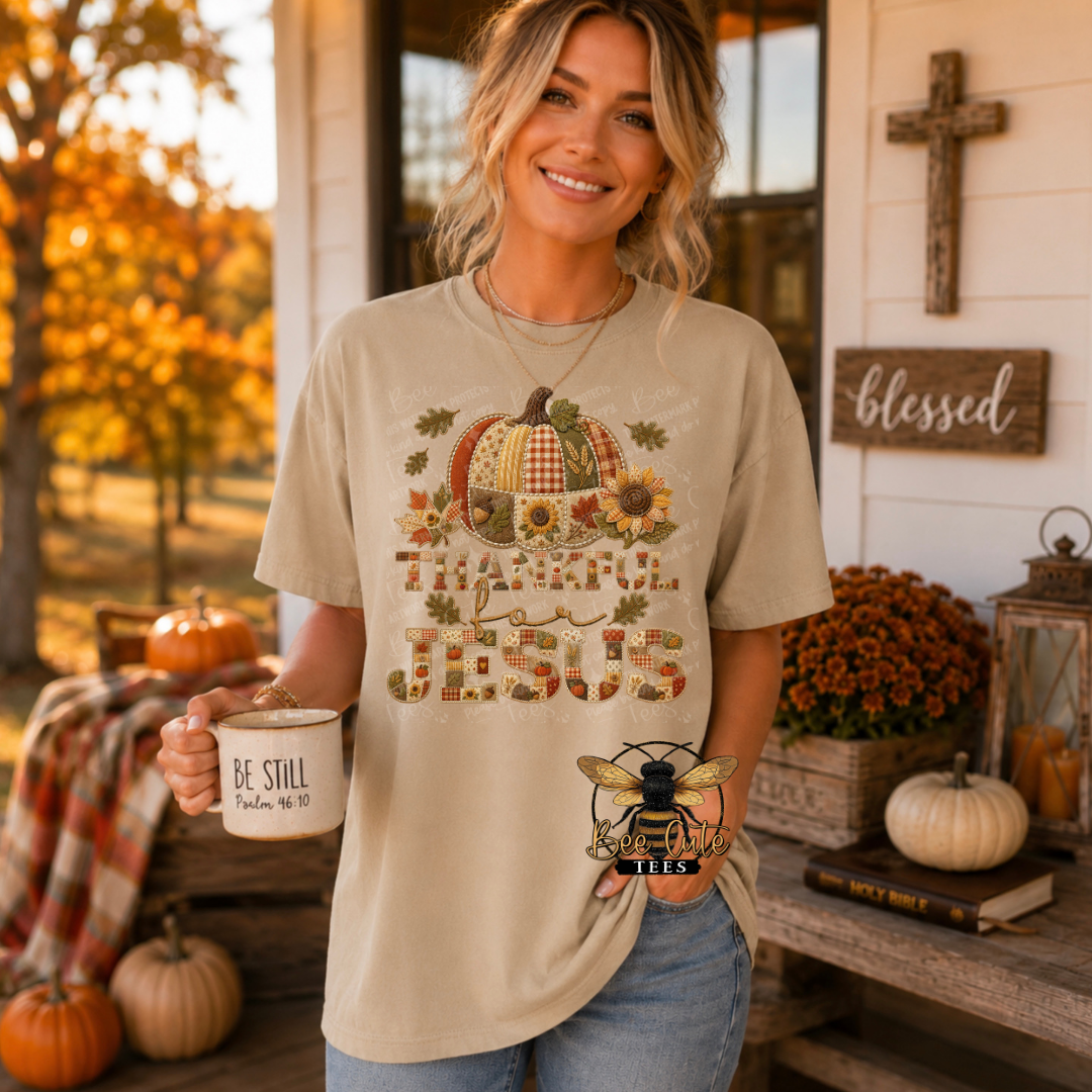 A woman stands on a decorated porch wearing a beige graphic T-shirt with a fall harvest design featuring pumpkins, sunflowers, leaves, and the words 'THANKFUL for JESUS.' She is holding a white mug that says 'BE STILL' and has a bee-shaped pin on her pocket.