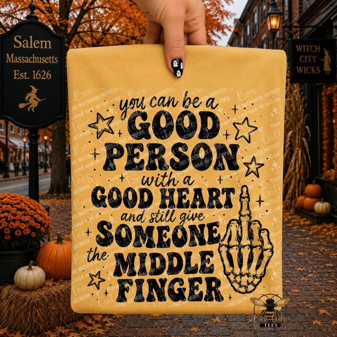 A hand holds a yellow t-shirt with black text that reads, 'you can be a GOOD PERSON with a GOOD HEART and still give SOMEONE the MIDDLE FINGER,' along with black star and skeleton hand graphics. The background shows a Halloween-themed street scene with pumpkins, autumn leaves, and storefront signs.