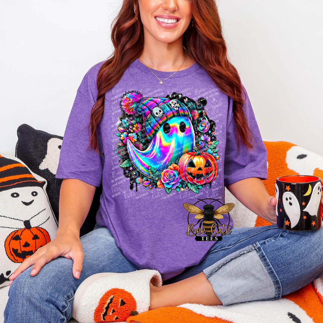 A woman wearing a purple graphic t-shirt with a colorful Halloween design featuring a ghost, jack-o'-lantern, and a bee logo. She is sitting on an orange and white blanket with Halloween-themed pillows and slippers, holding a black mug with a ghost design.