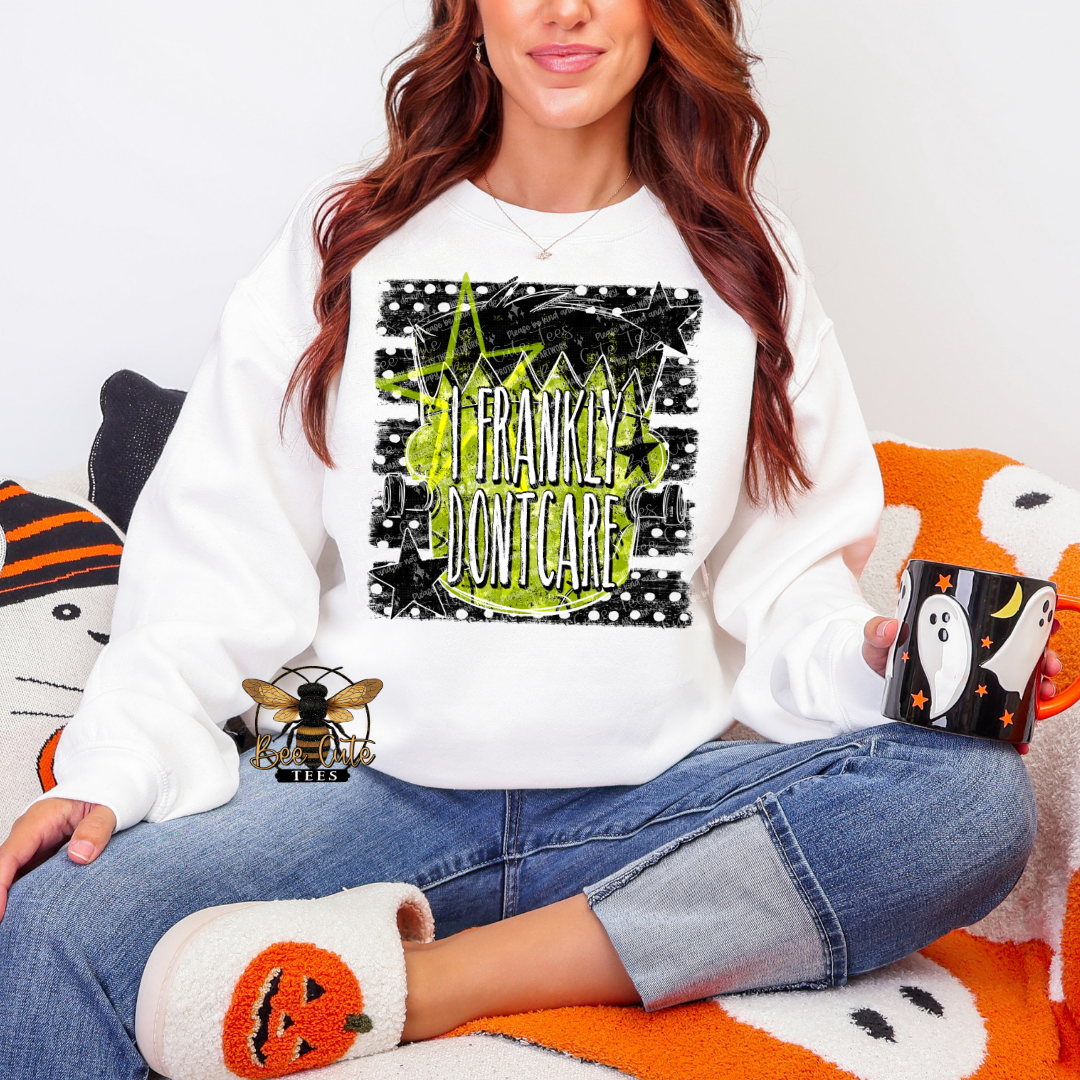 A woman wearing a white crewneck sweatshirt with a black and lime green 'I FRANKLY DON'T CARE' Halloween graphic, paired with blue jeans and white slippers, sitting on an orange and white blanket.