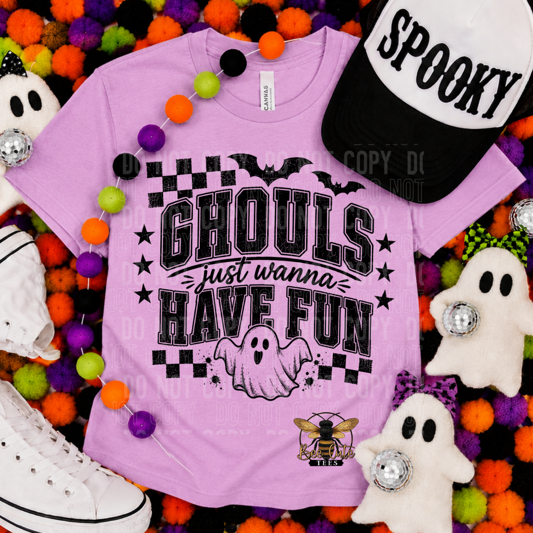 A lavender purple graphic t-shirt with a black 'Ghouls just wanna HAVE FUN' design, styled in a flat-lay with Halloween props including a 'Spooky' cap, white plush ghosts, colorful pom-pom garlands, and white sneakers.