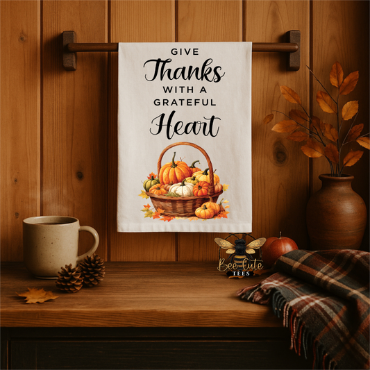 A white kitchen towel with a black script quote reading 'GIVE Thanks WITH A GRATEFUL Heart' and an autumn harvest illustration of pumpkins, hanging on a wooden wall rack in a cozy fall-themed setting with a mug, pinecones, a plaid throw, and a vase of leaves.
