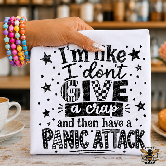 A hand wearing colorful beaded bracelets holds a white t-shirt with black text that reads, 'I'm like I don't GIVE a crap and then have a PANIC ATTACK.' The sign is photographed on a wooden table in a cafe-like setting with a coffee cup and pastries blurred in the background.