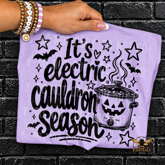 A hand holds a lavender purple t-shirt featuring a black graphic that reads 'It's electric Cauldron Season' with a jack-o'-lantern slow cooker, stars, and bats against a black brick wall background.