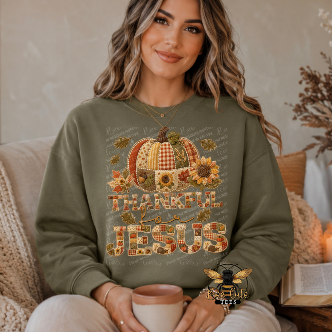 A woman sitting indoors wearing an olive green sweatshirt with a large autumn-themed graphic that reads 'THANKFUL JESUS' and features a pumpkin, sunflowers, leaves, and patterned lettering. She is holding a mug in a cozy room setting.