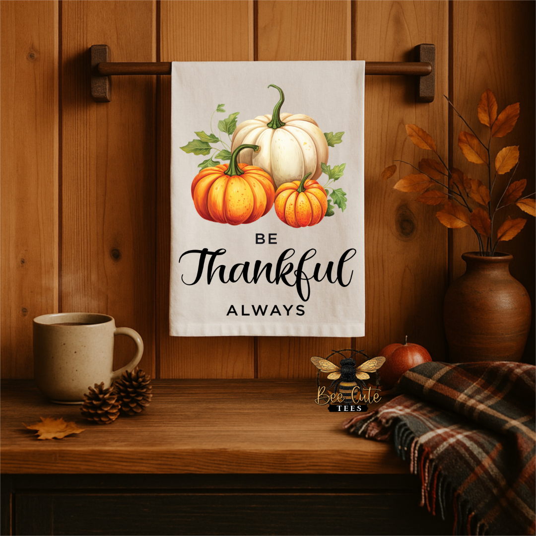 A white kitchen towel with a fall harvest design hangs on a wooden rack. The towel features three pumpkins (two orange, one white) with green leaves and stems, above the text 'BE Thankful ALWAYS' in black script.