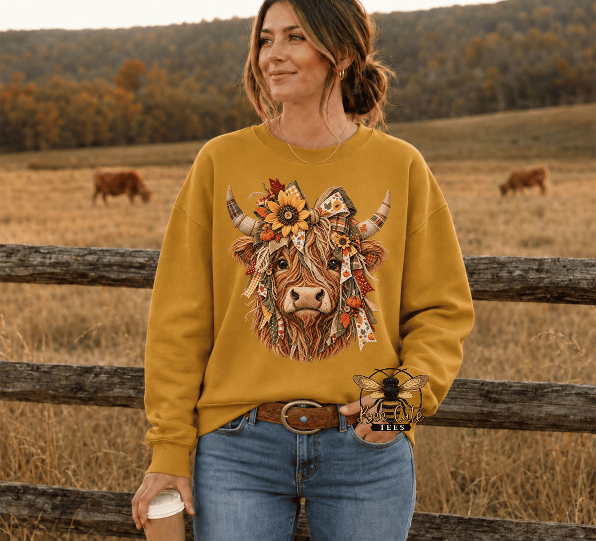 A woman wearing a mustard yellow crewneck sweatshirt with a detailed graphic of a cow decorated with flowers and a cowboy hat, set against a rustic outdoor background with a wooden fence and grazing cows.