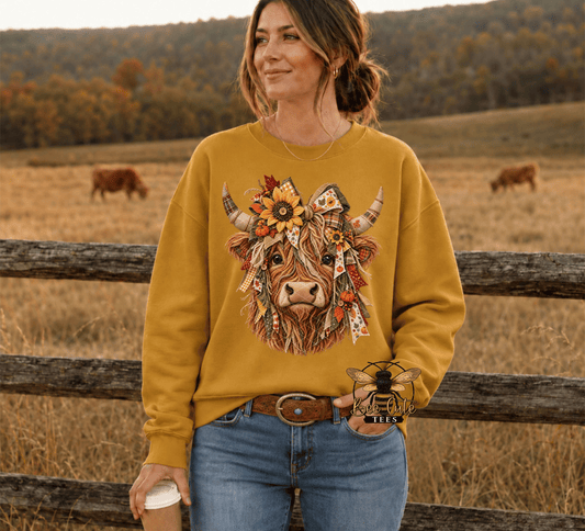 A woman wearing a mustard yellow crewneck sweatshirt with a detailed graphic of a cow decorated with flowers and a cowboy hat, set against a rustic outdoor background with a wooden fence and grazing cows.