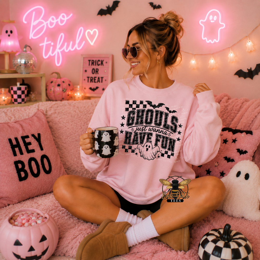 A woman sits cross-legged on a pink fluffy couch wearing a light pink graphic sweatshirt that reads 'Ghouls just wanna have fun' with a ghost and checkerboard design. She holds a black mug with white ghosts, surrounded by Halloween decorations including a 'Hey Boo' pillow, jack-o'-lanterns, and glowing neon signs.