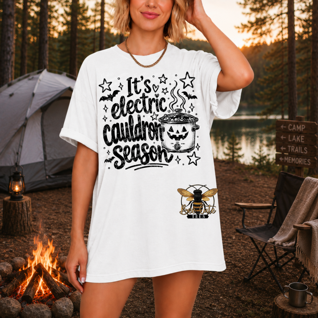 A woman modeling a white graphic T-shirt in an outdoor campsite setting at sunset, with a tent, campfire, lake, and trees in the background. The shirt features black text reading 'It's electric cauldron season' with a cauldron graphic and a bee illustration near the hem.