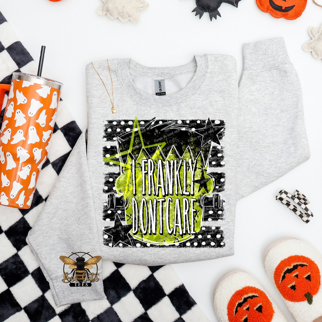 A heather gray crewneck sweatshirt with a Halloween-themed graphic print reading 'Frankly Don't Care' in green and black. The sweatshirt is styled in a flat-lay arrangement with a black and white checkered blanket, a ghost-patterned tumbler, a bee applique patch, and pumpkin-themed slippers on a white background.