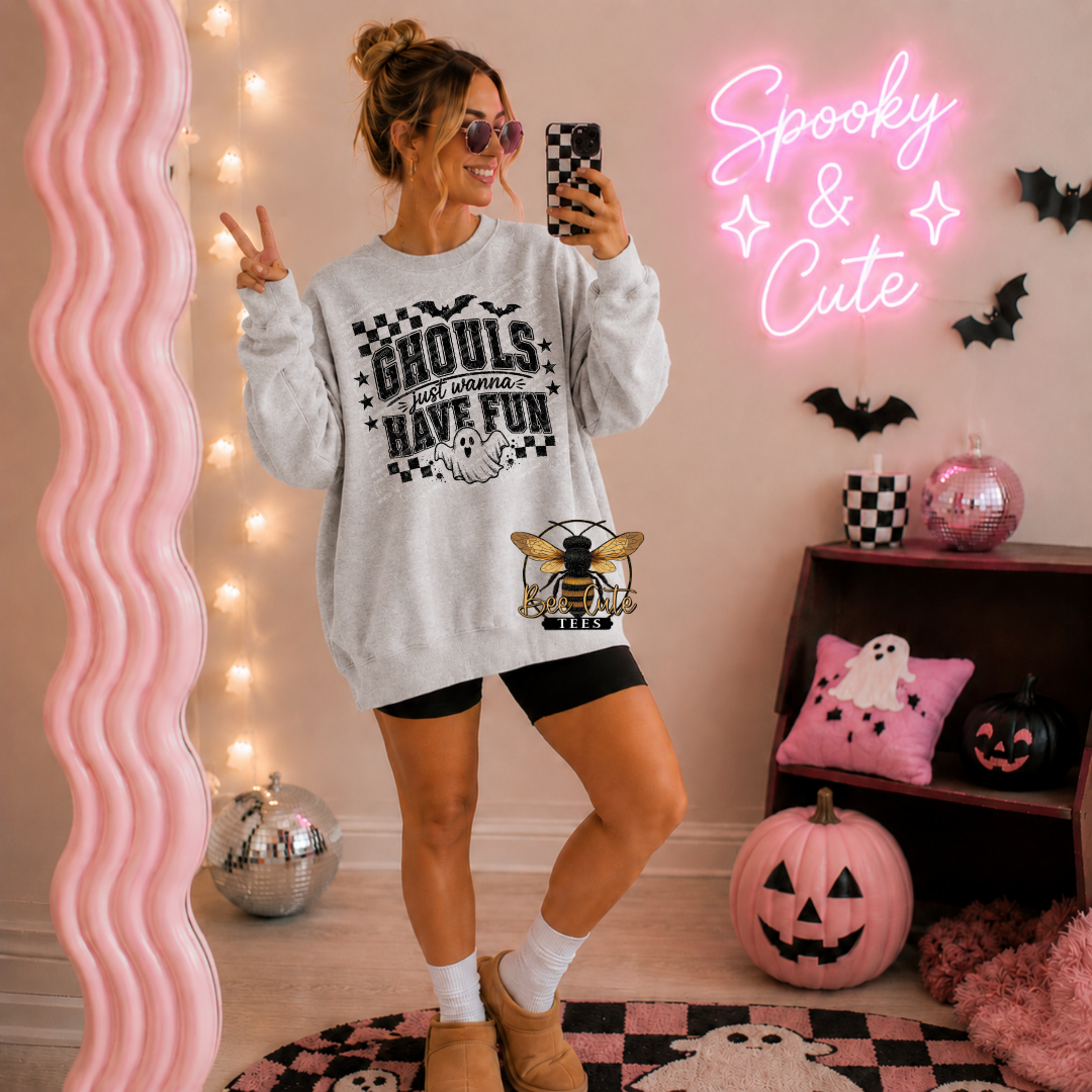 A woman posing in a cozy, Halloween-themed room wearing a light gray crewneck sweatshirt with a black graphic that reads 'Ghouls that always have fun' and a bee design. The room features pink decor, a pink wavy mirror, a pink neon sign reading 'Spooky & Cute', and pumpkins on a shelf.