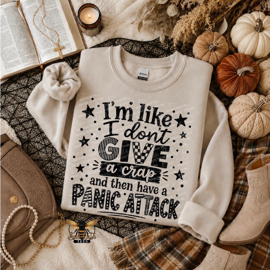 A cream-colored crewneck sweatshirt with a black graphic text print reading 'I'm like I don't GIVE a crap and then have a PANIC ATTACK' styled in a cozy autumn flat-lay with pumpkins, boots, a plaid scarf, and an open book.
