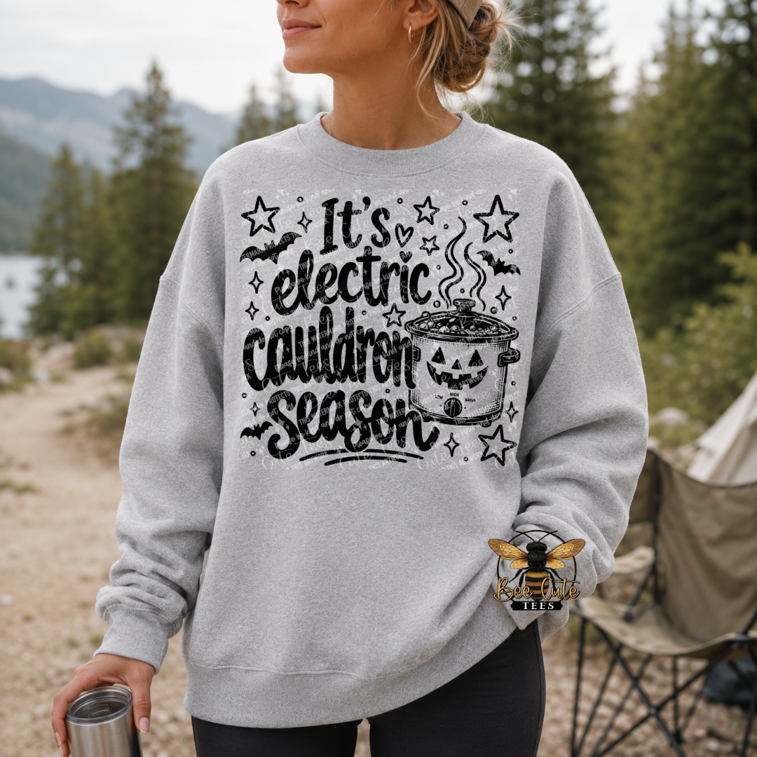 A woman wearing a light gray crewneck sweatshirt with a black graphic that reads 'It's electric cauldron Season' surrounded by stars, bats, and a cauldron illustration. She is standing outdoors in a wooded setting near a tent.