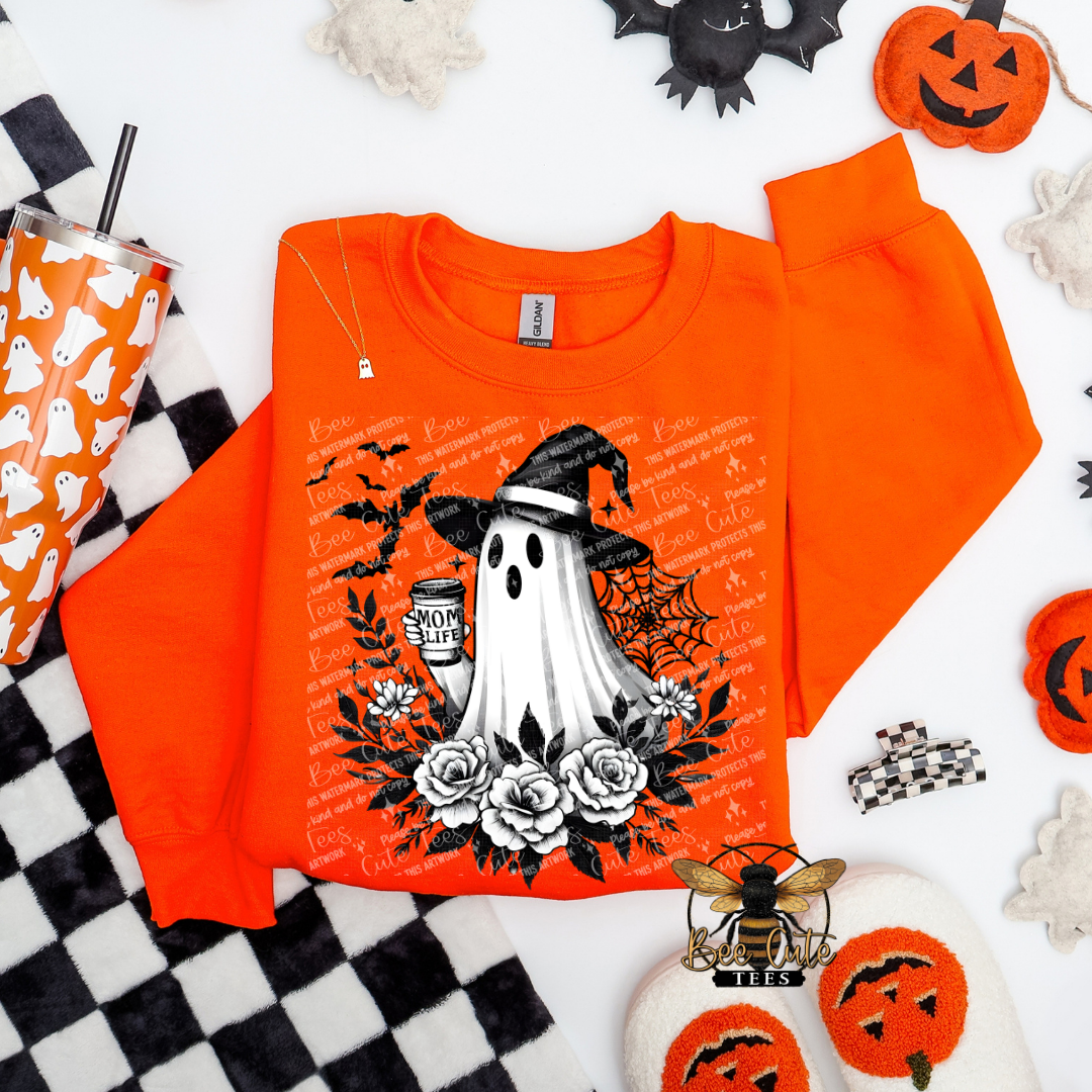 An orange crewneck sweatshirt with a Halloween-themed graphic print featuring a ghost, witch hat, bats, roses, and a cup, styled in a flat lay with pumpkin decorations, a checkered blanket, and a patterned tumbler.