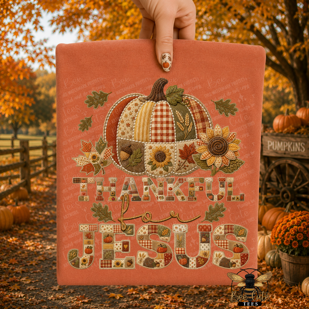 A hand holds a folded t-shirt featuring a patchwork pumpkin, autumn leaves, sunflowers, and the words “THANKFUL JESUS” in applique-style lettering. The sign has a warm peach background and is photographed outdoors in a fall yard with pumpkins, orange foliage, and a wooden fence visible.