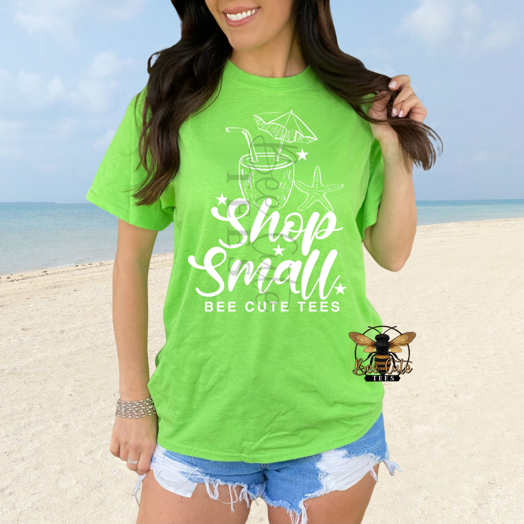 A woman wearing a bright lime green graphic t-shirt with the text 'Shop Small' and 'BEE CUTE TEES' in white script, standing on a sandy beach with the ocean in the background.