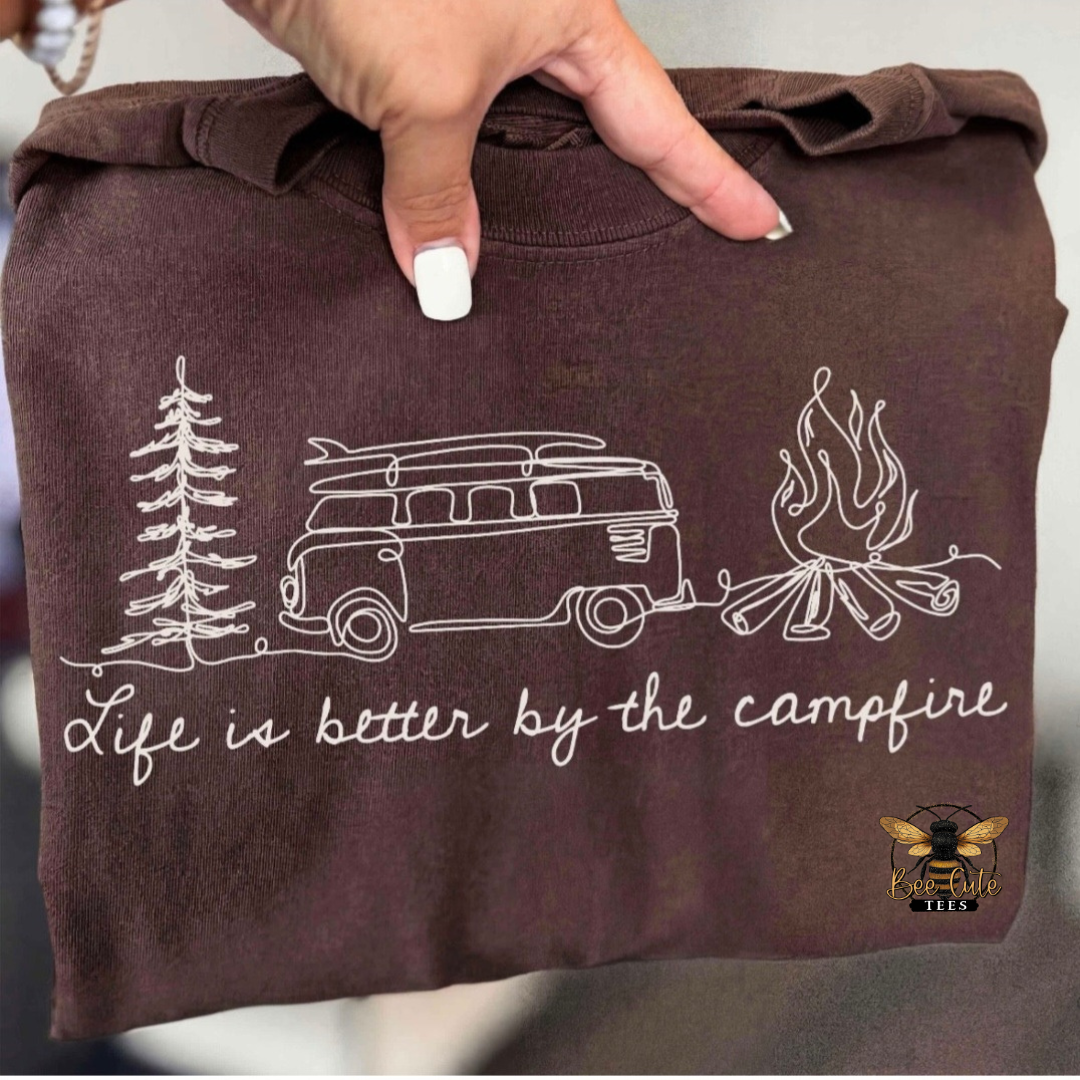 A folded brown sweatshirt held up by a hand, featuring a white line art graphic of a camp van, trees, campfire, and the text 'Life is better by the campfire'. A small bee logo in the bottom right corner.