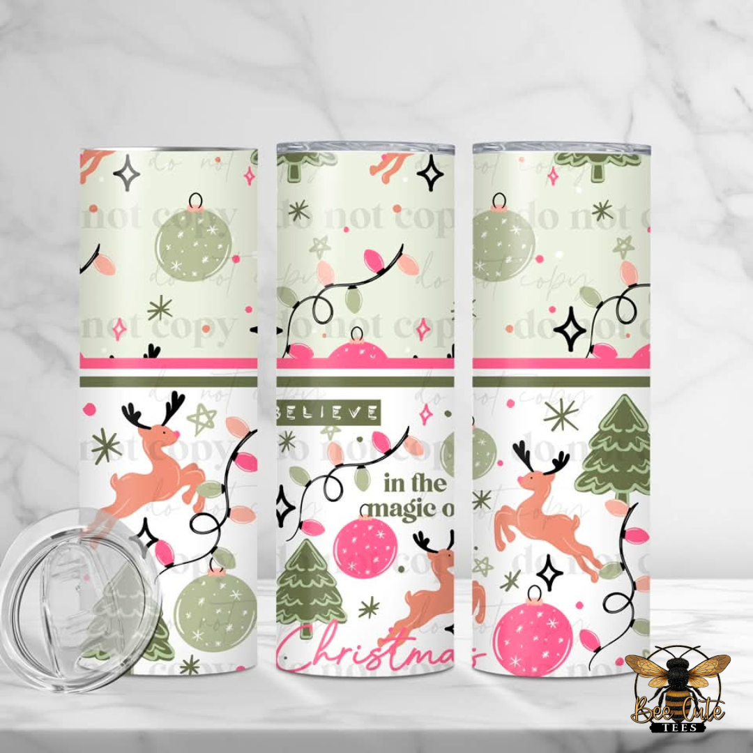 Three insulated tumbler cups with a festive Christmas pattern featuring reindeer, ornaments, trees, and text, displayed on a light marble surface with a clear lid in the foreground.