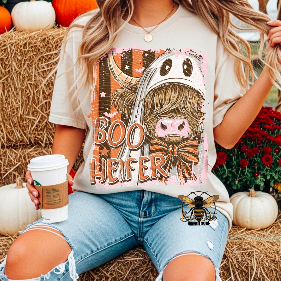 A woman wearing a graphic t-shirt with a 'Boo Heifer' cartoon design, sitting in a fall-themed setting with hay bales, pumpkins, and flowers.