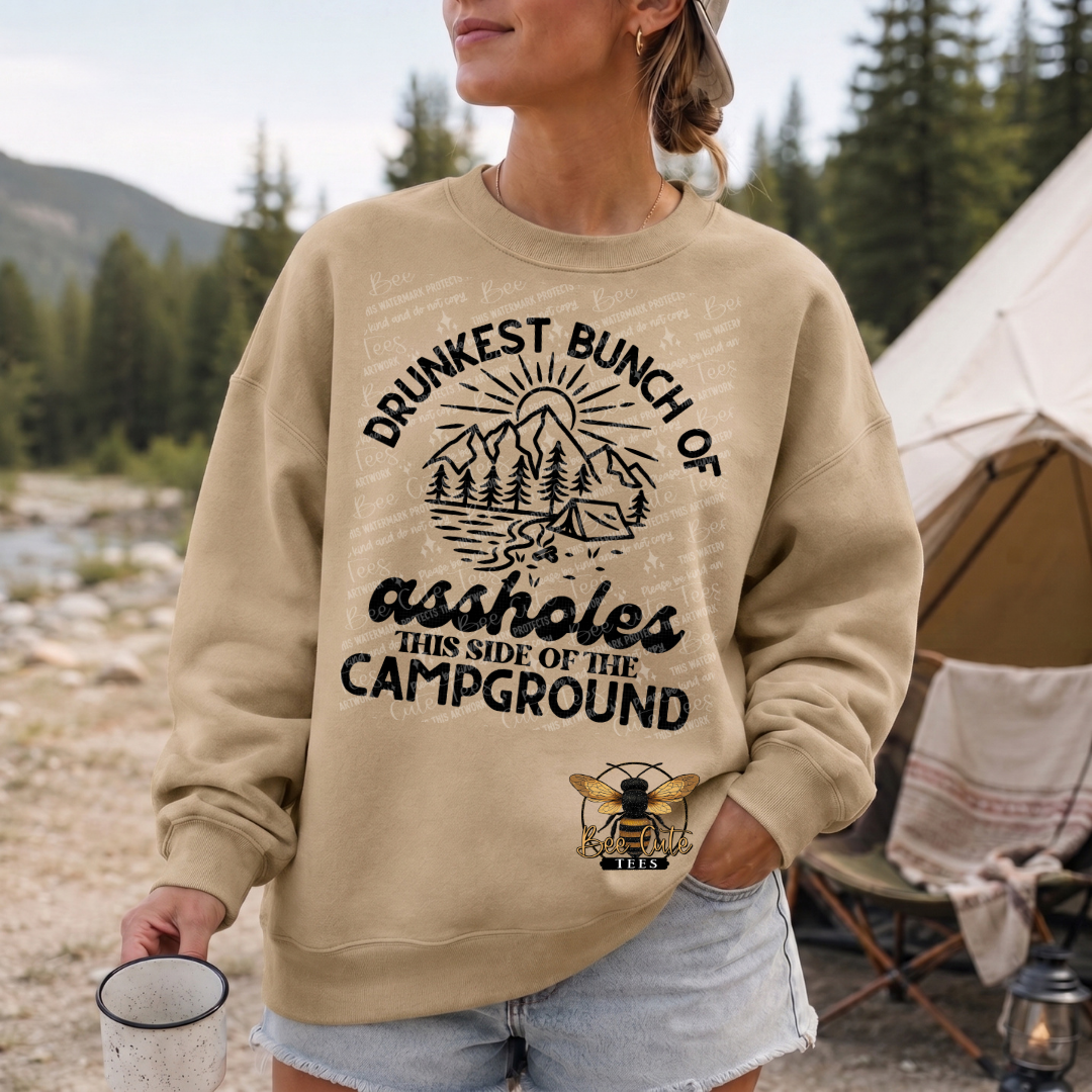 A woman wearing a beige crewneck sweatshirt with a black graphic that reads 'DRUNKEST BUNCH OF ASSHOLES THIS SIDE OF THE CAMPGROUND' and a bee logo, standing outdoors near a tent and trees.