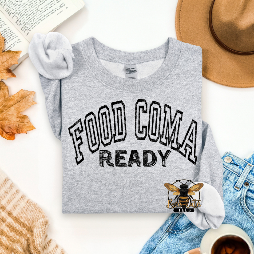 A folded heather gray graphic T-shirt with the words 'FOOD COMA READY' printed in black outlined lettering, styled in a flat lay with a hat, jeans, knit sweater, leaves, an open book, and a cup on a white background.