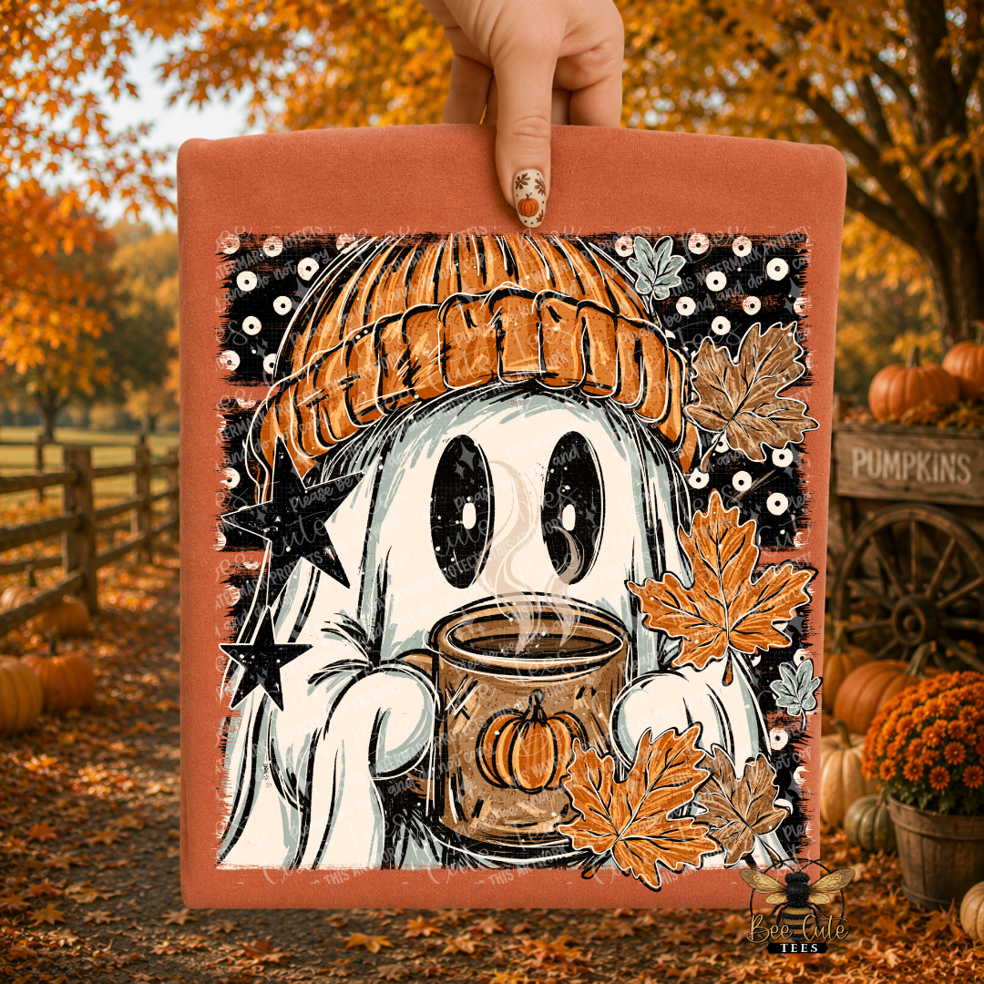 A hand holds a folded orange t-shirt featuring a cartoon ghost in an orange knit beanie holding a mug, surrounded by autumn leaves and pumpkins, with an outdoor fall yard.