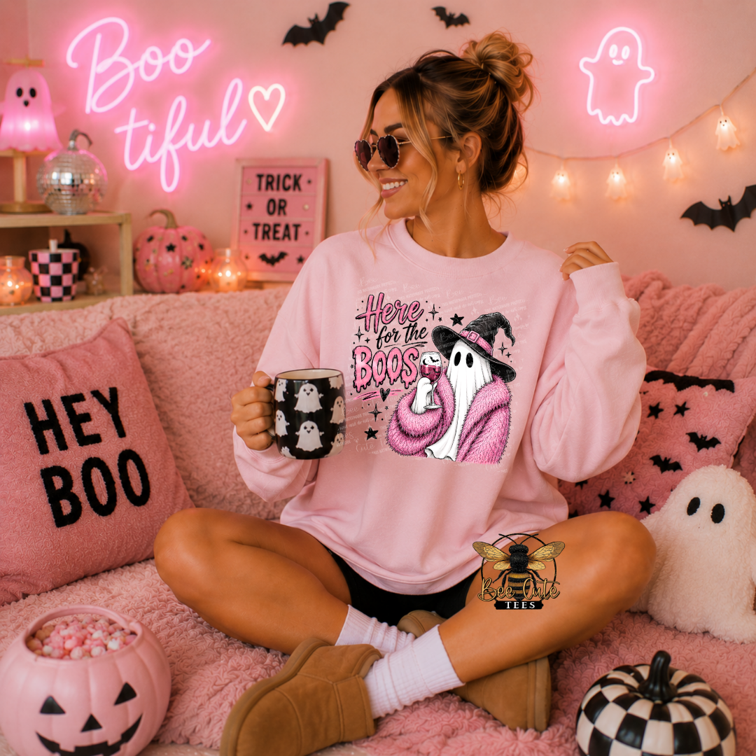 A woman sits on a pink couch wearing a light pink graphic sweatshirt with a cartoon ghost and the text 'Here for the Boos'. She holds a mug with ghost illustrations, surrounded by Halloween decorations including a 'HEY BOO' pillow, jack-o'-lanterns, and a 'Boo Tiful' neon sign.