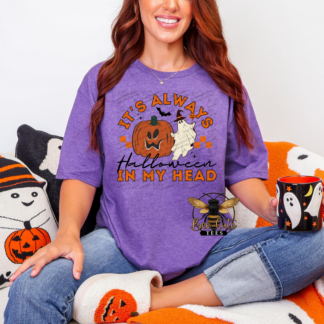 A woman wearing a purple graphic t-shirt with a Halloween design that reads 'IT'S ALWAYS Halloween IN MY HEAD' with a jack-o'-lantern, ghost, and bee logo, sitting on an orange and white blanket with Halloween-themed pillows.