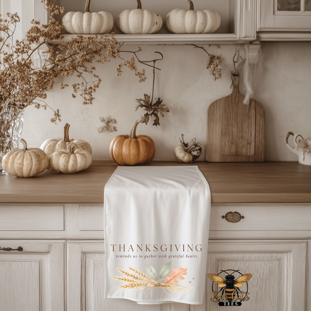 A white Thanksgiving kitchen tea towel draped over a wooden kitchen cabinet, featuring the text 'THANKSGIVING' and a wheat and leaf illustration. The scene is decorated with pumpkins, a wooden cutting board, and dried floral arrangements.