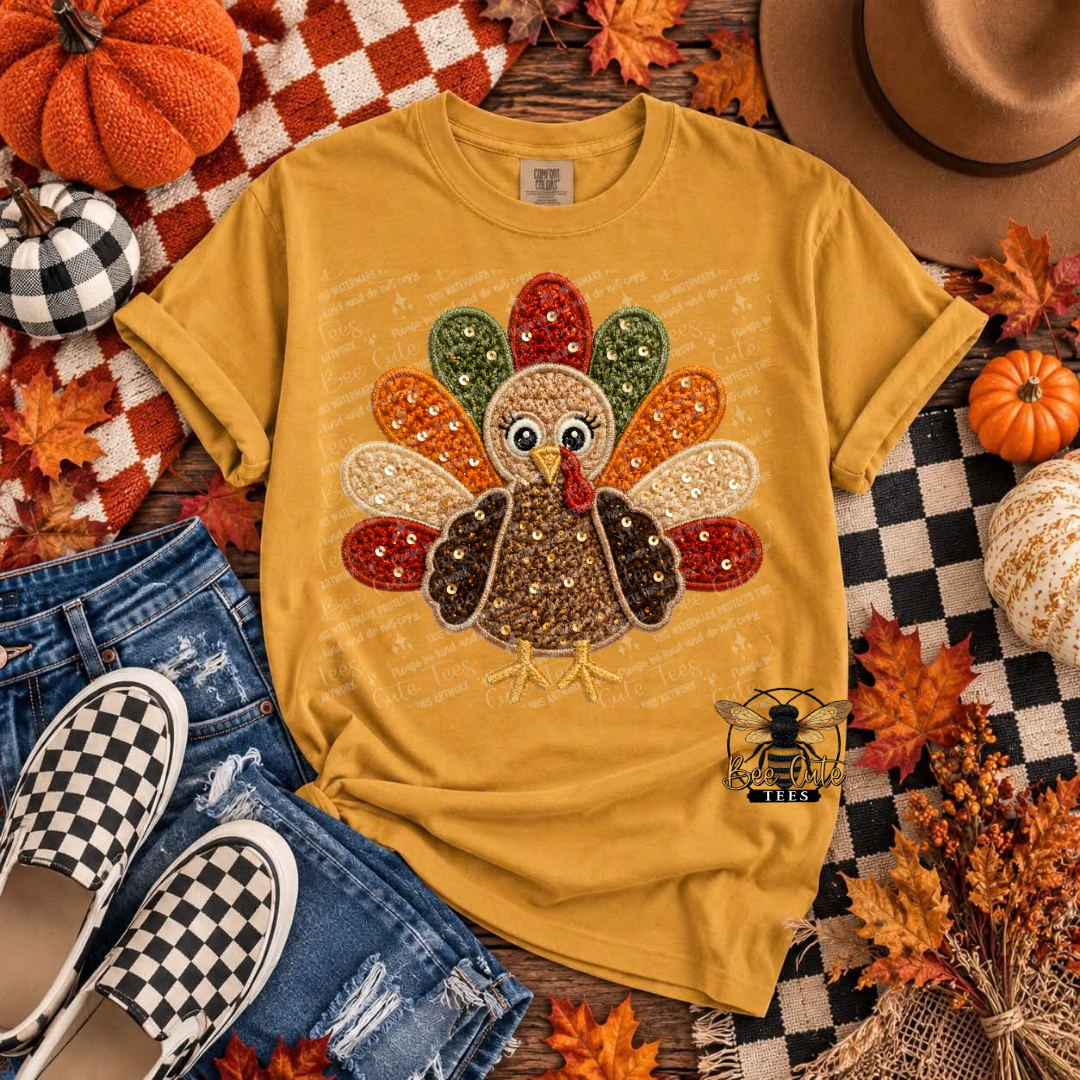 A mustard yellow graphic t-shirt featuring a sequined turkey design, styled in a flat lay with autumn props including pumpkins, a hat, jeans, and checkered shoes.