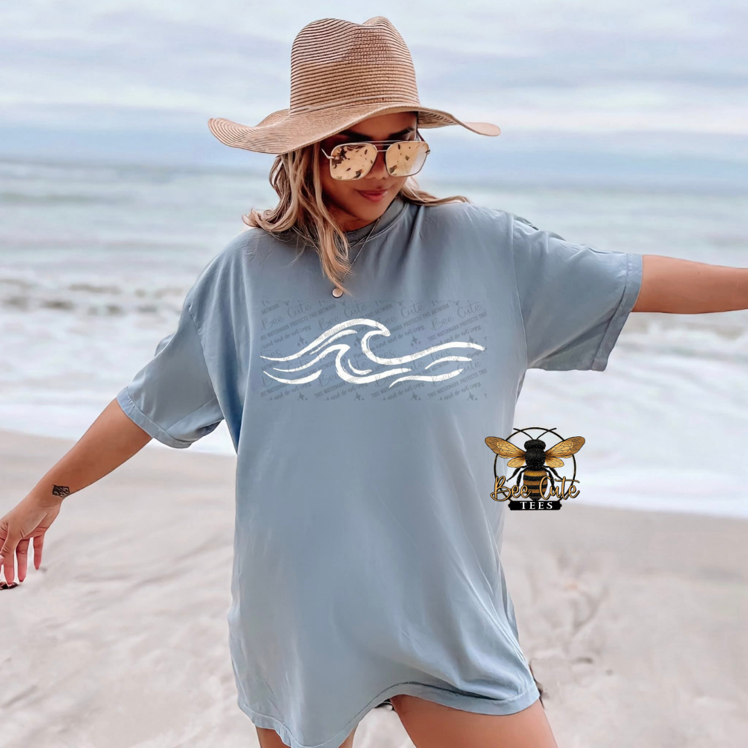 Woman on a beach wearing a light blue t-shirt with a wave design, sunglasses, and a wide-brimmed hat.