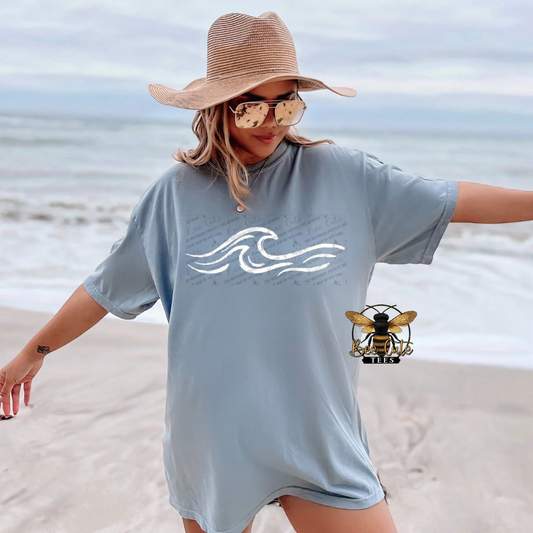 Woman on a beach wearing a light blue t-shirt with a wave design, sunglasses, and a wide-brimmed hat.