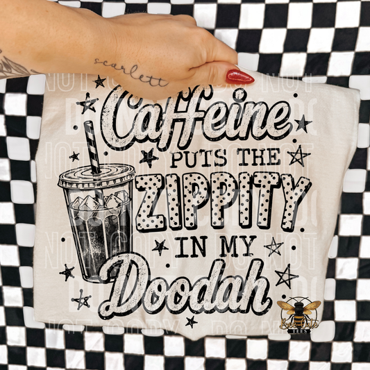 A hand with red nail polish holds a cream-colored t-shirt featuring a black and white graphic that reads 'Caffeine PUTS THE ZIPPITY IN MY Doodah' with a coffee cup illustration and a bee logo. The background is a black and white checkered pattern.
