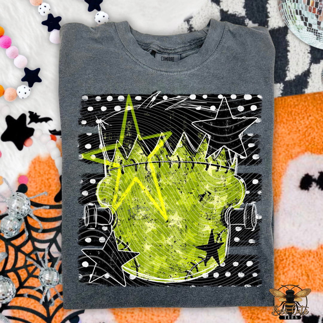 A folded gray crewneck t-shirt with a distressed lime green and black graphic print, laid flat on a Halloween-themed background with candy, spiderwebs, and a disco ball.