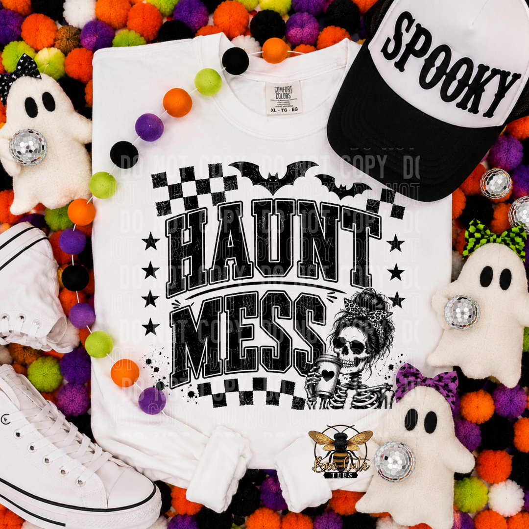 A white graphic T-shirt laid flat with a Halloween-themed design reading 'HAUNT MESS' in black block letters, with bats, stars, checkerboard borders, and a skeleton illustration. The shirt is styled in a flat lay with a black-and-white 'SPOOKY' cap, plush ghosts, pom-pom garland, white sneakers, and small decorative accessories around it.
