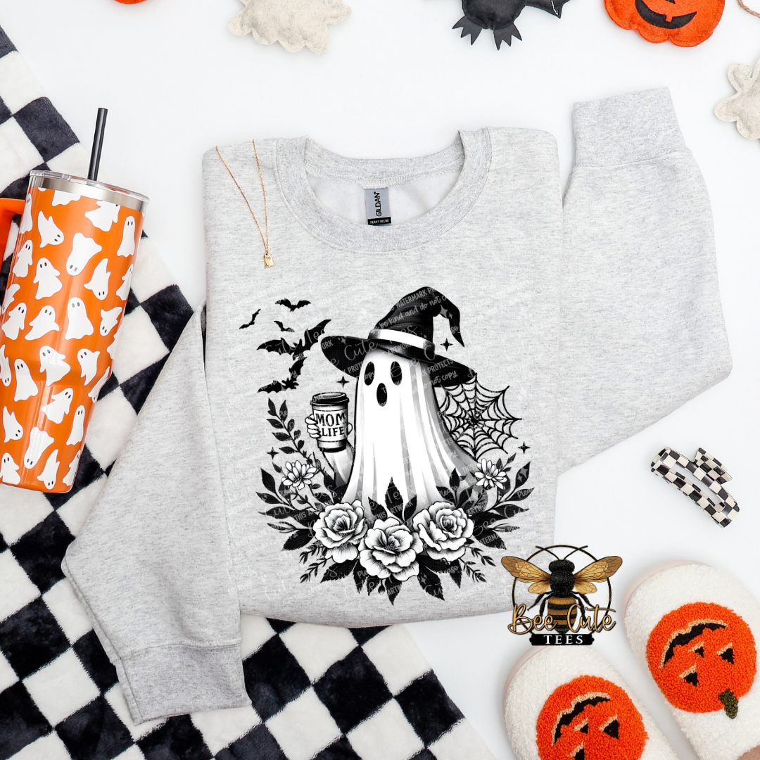 A light heather gray sweatshirt laid flat on a white background with Halloween-themed props, featuring a black graphic of a ghost in a witch hat surrounded by roses, bats, a spiderweb, and a mug.