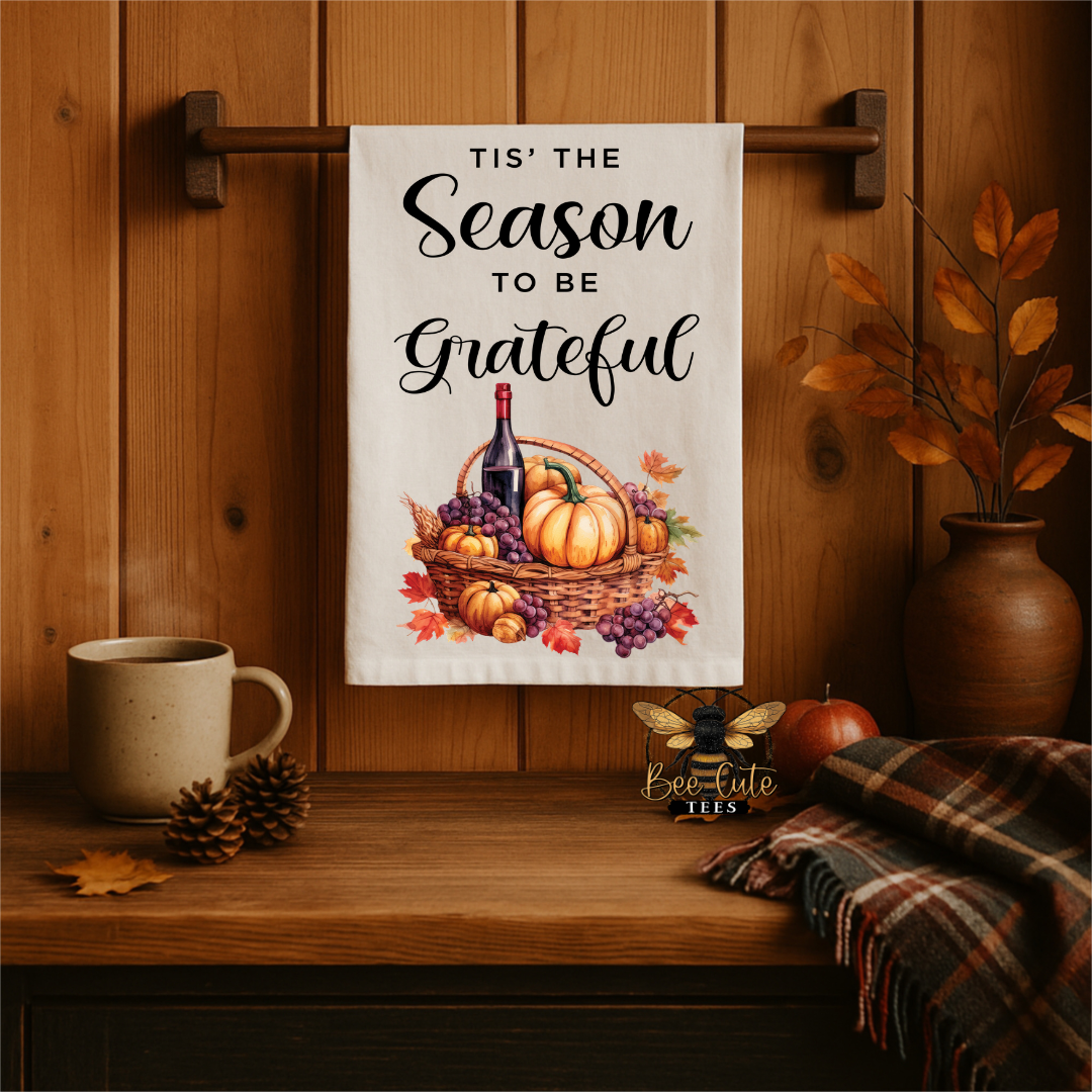 A white kitchen towel with a fall-themed illustration and the text 'TIS' THE Season TO BE Grateful' hangs on a wooden wall rack. The scene is styled with a ceramic mug, pinecones, a plaid throw blanket, a small apple, and a vase of autumn leaves on a wooden table.