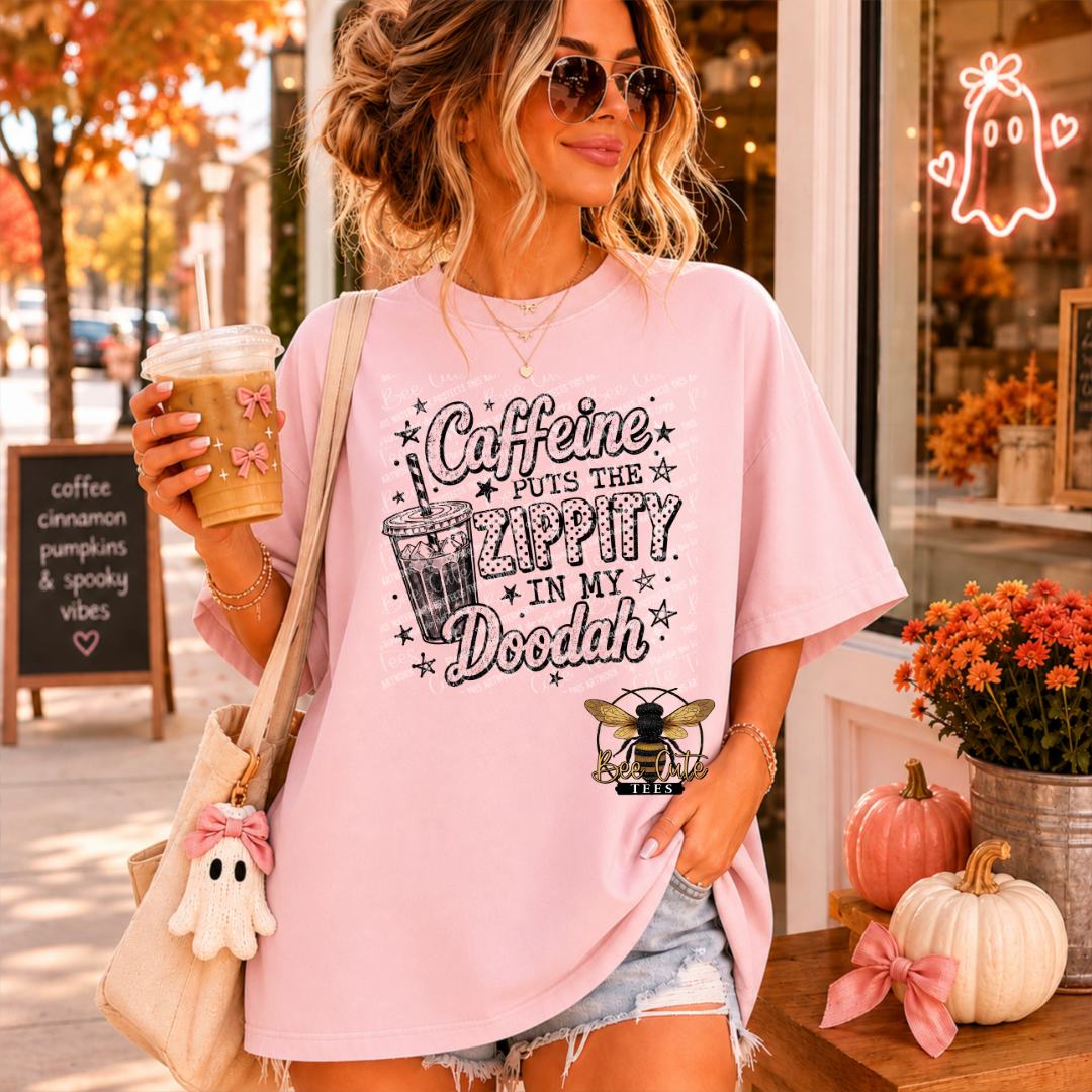 A woman wearing a light pink graphic t-shirt with a 'Caffeine PUTS THE ZAPPY IN MY DOODAH' design and a bee logo, holding a coffee cup in a storefront setting.