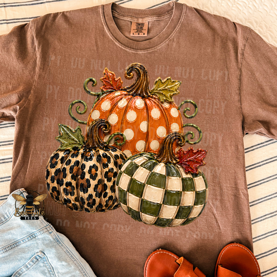 A brown short-sleeve t-shirt laid flat on a striped fabric background, featuring a large graphic of three decorative pumpkins in orange, leopard print, and green-and-white checkered patterns. Light blue jeans and brown sandals are partially visible as styling props.