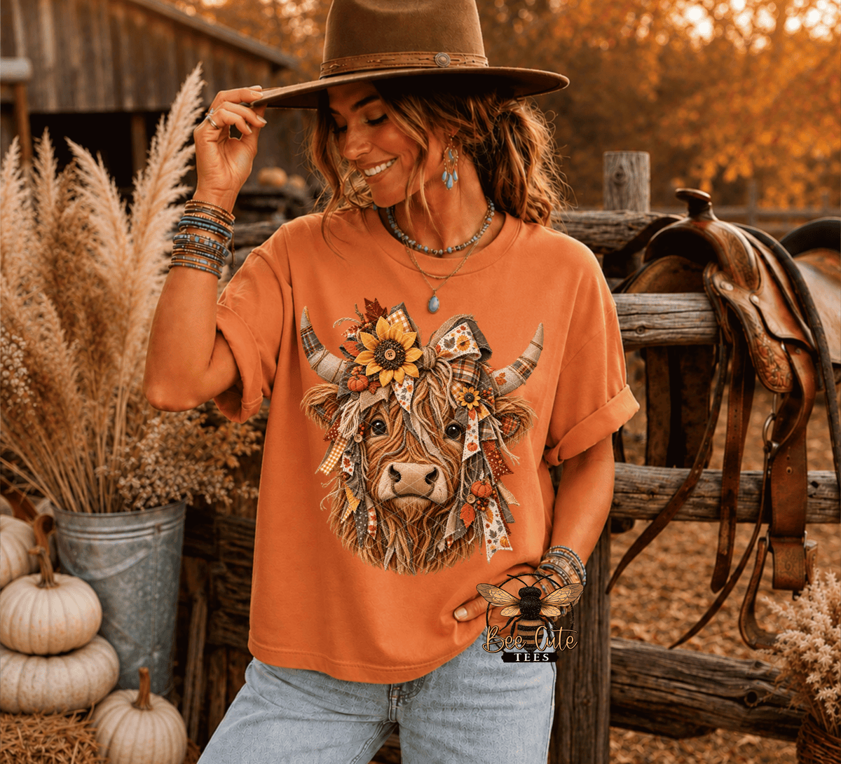 A woman wearing a rust-orange graphic t-shirt with a detailed Highland cow design, styled with a brown wide-brim hat, bracelets, and light-wash jeans in a rustic outdoor setting with a wooden fence, saddle, and pumpkins.