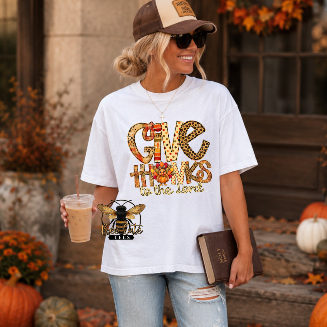 A woman wearing a white graphic t-shirt with a 'Give Thanks to the Lord' autumn-themed design, styled with a brown and beige baseball cap, sunglasses, and jeans. She is holding an iced coffee and a book, standing in front of a rustic autumn porch decorated with pumpkins and fall foliage.