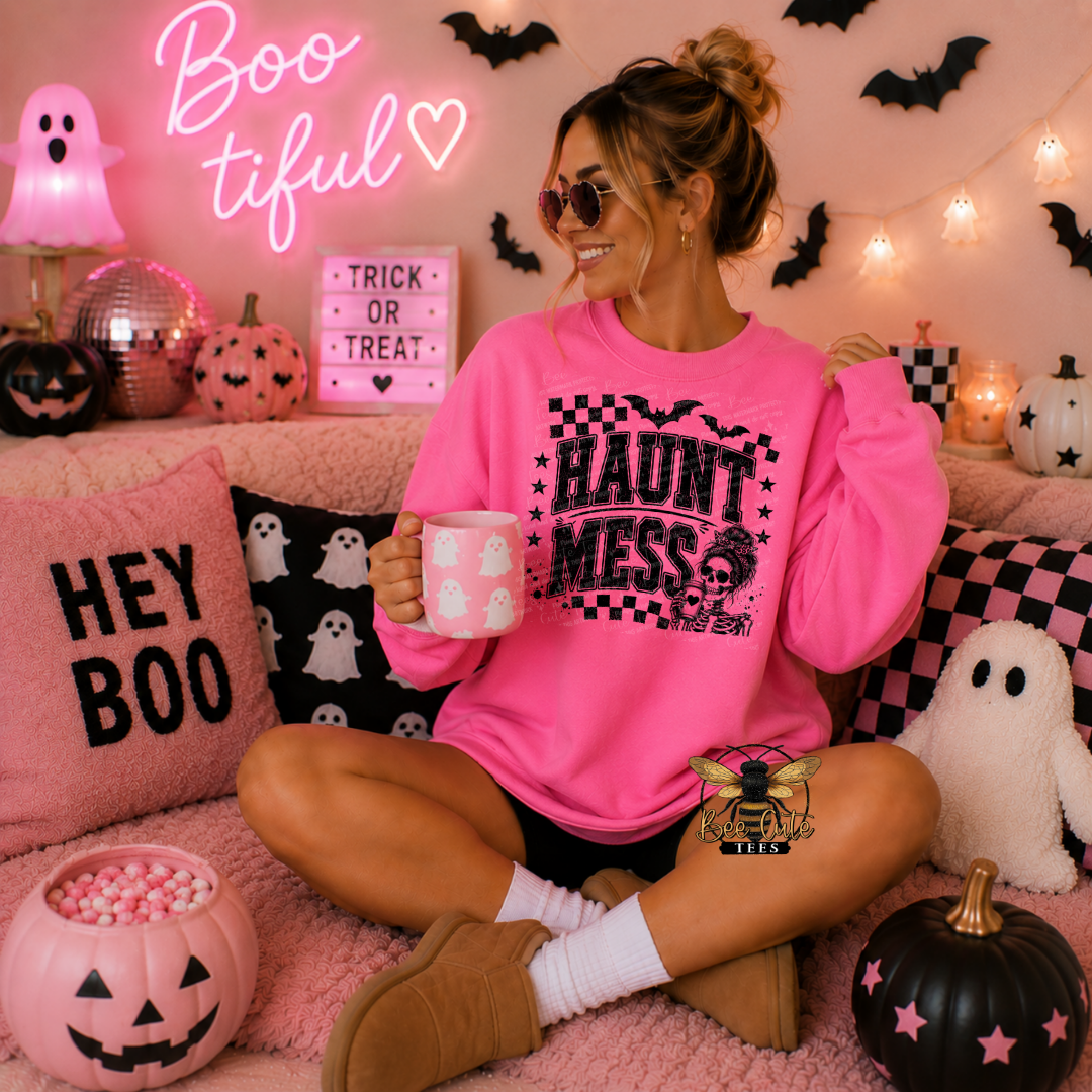 A woman sits cross-legged on a pink couch wearing a bright pink sweatshirt with a 'HAUNT MESS' graphic, surrounded by Halloween decorations including a jack-o'-lantern, ghost pillows, and a pink neon sign.
