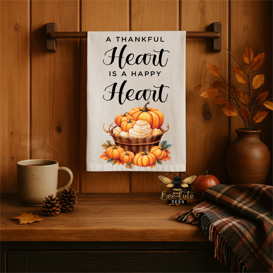 A white kitchen towel with the phrase 'A THANKFUL Heart IS A HAPPY Heart' in black script, featuring an illustration of orange pumpkins and a basket of white pumpkins, hanging on a wooden wall rack in a cozy autumn-themed setting with a mug, pinecones, and a plaid throw blanket.