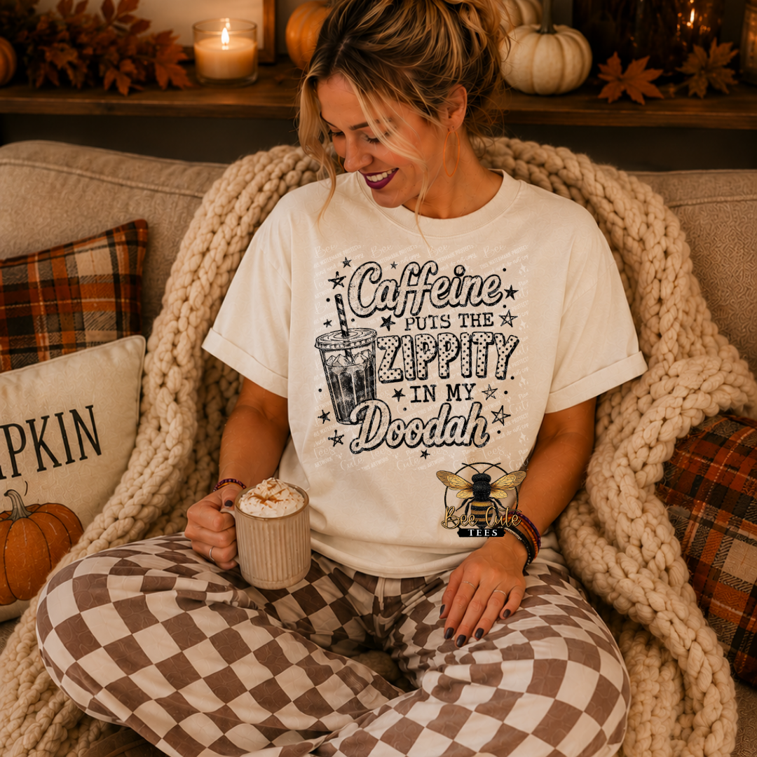 A woman sitting on a couch wrapped in a chunky knit blanket, wearing a white graphic t-shirt that reads 'Caffeine PUTS THE ZAPPY IN MY Doodah' with a coffee cup and bee logo, paired with brown and white checkered pajama pants.
