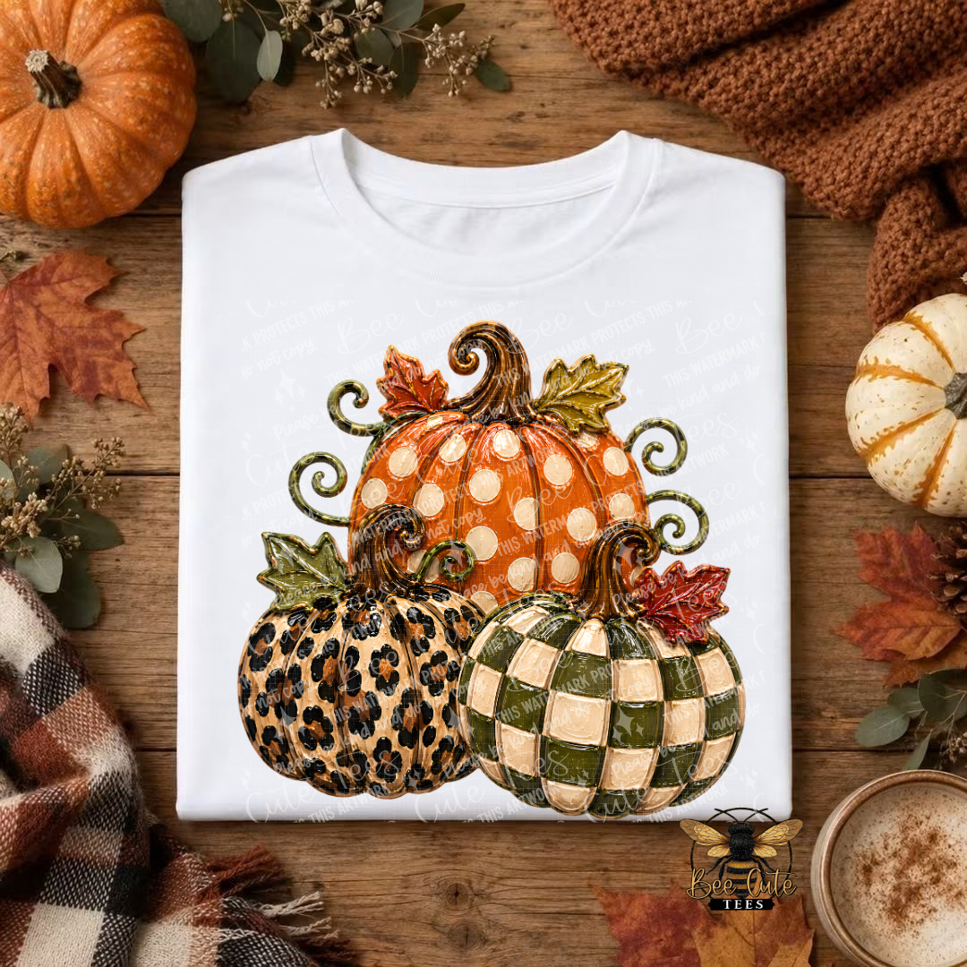 A folded white T-shirt with a large autumn pumpkin graphic, displayed on a rustic wooden surface surrounded by fall leaves, a small orange pumpkin, a knit brown sweater, and a cup of coffee. The graphic features three pumpkins: one orange with white polka dots, one leopard print, and one green-and-beige checkered pattern.