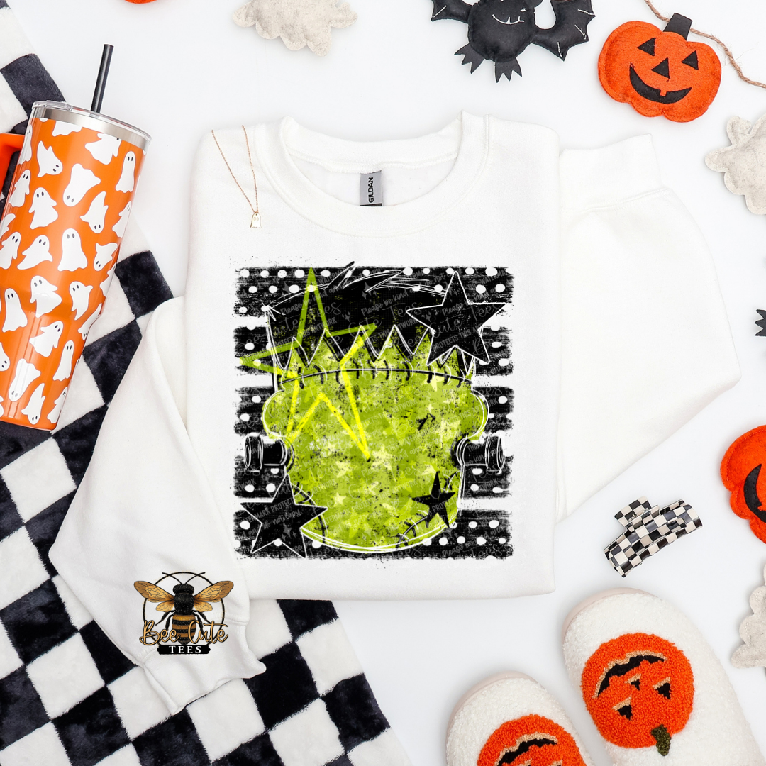 A white long-sleeve crewneck sweatshirt with a Halloween-themed graphic print of a green cauldron, black stars, and a polka-dot border, styled in a flat lay with seasonal props including a ghost-patterned tumbler, black-and-white checkered blanket, and orange jack-o'-lantern decorations.