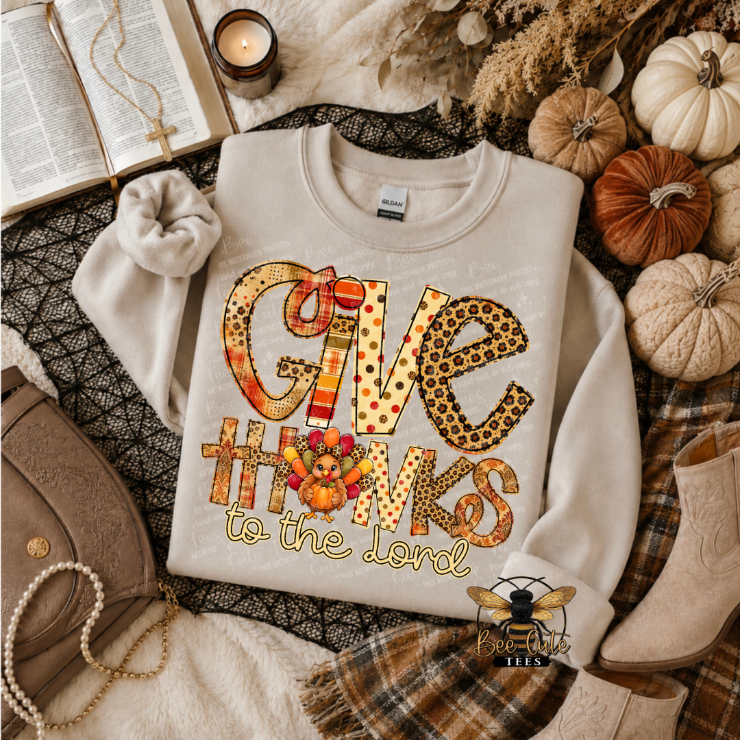 A folded cream sweatshirt with a large 'GIVE THANKS to the Lord' autumn-themed graphic, styled in a cozy flat lay with pumpkins, a book, a candle, a handbag, and plaid fabric.